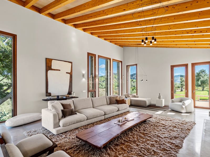 Salon lumineux avec hauts plafonds en bois, canapé crème, table en bois massif et grandes baies vitrées.