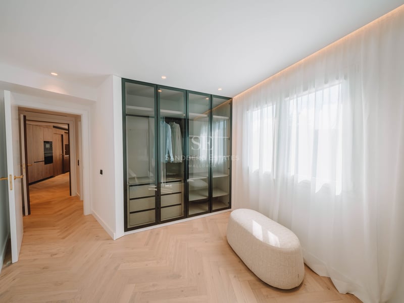 Dressing moderne avec parquet en chevrons, armoire vitrée et pouf central près d'une fenêtre.