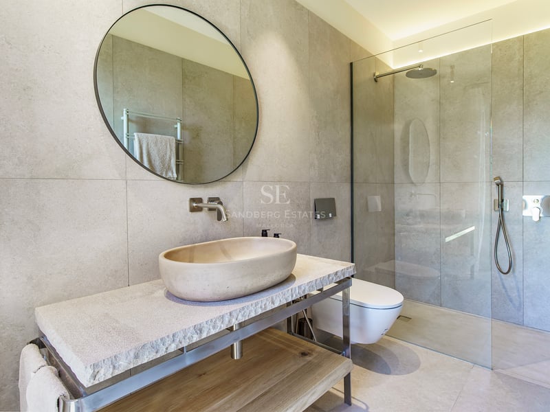 Modernes Badezimmer mit ovalem Steinbecken, Rundspiegel und begehbarer Glasdusche mit großen Fliesen.