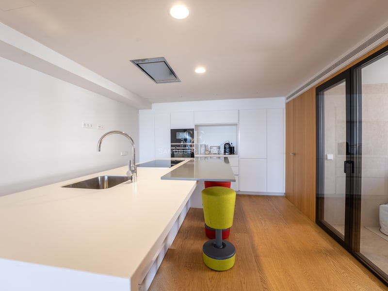 Ljust modernt kök med en stor vit köksö, diskho, trägolv och inbyggda vitvaror.