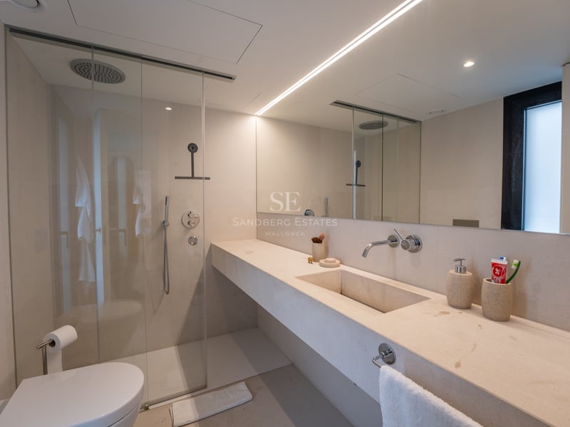 Modernt badrum med handfat i beige sten, duschvägg i glas, stor spegel och linjär belysning.