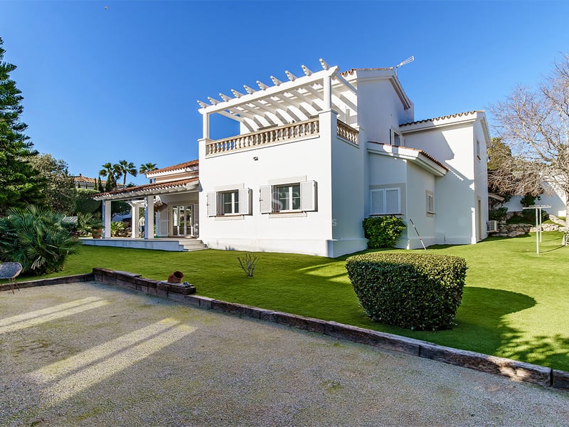 Vit tvåplansvilla med pergola på taket, välskött gräsmatta och pinjeträd mot en klarblå himmel.