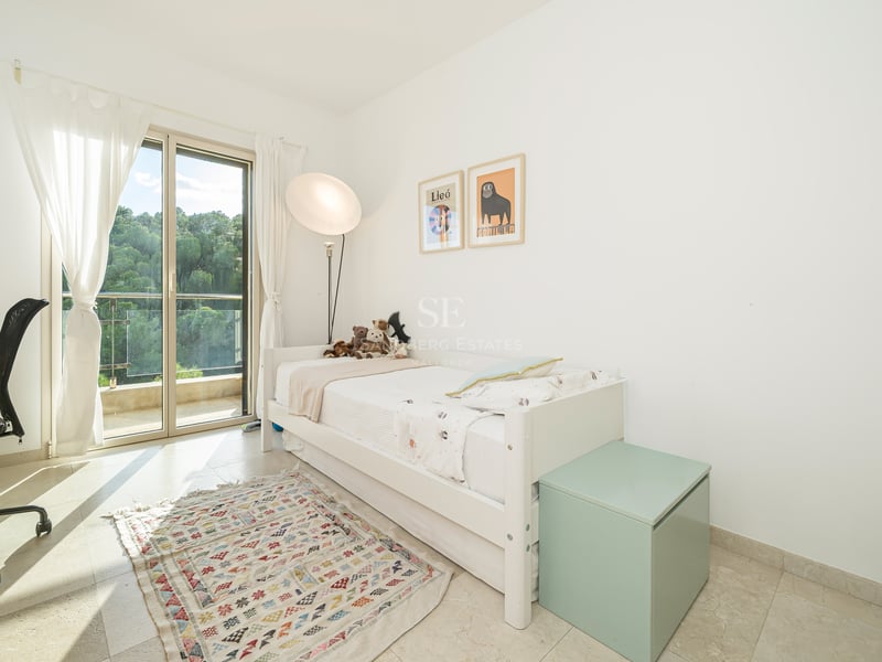 Vitt sovrum med enkelsäng, skrivbord, balkongdörr och mönstrad matta.