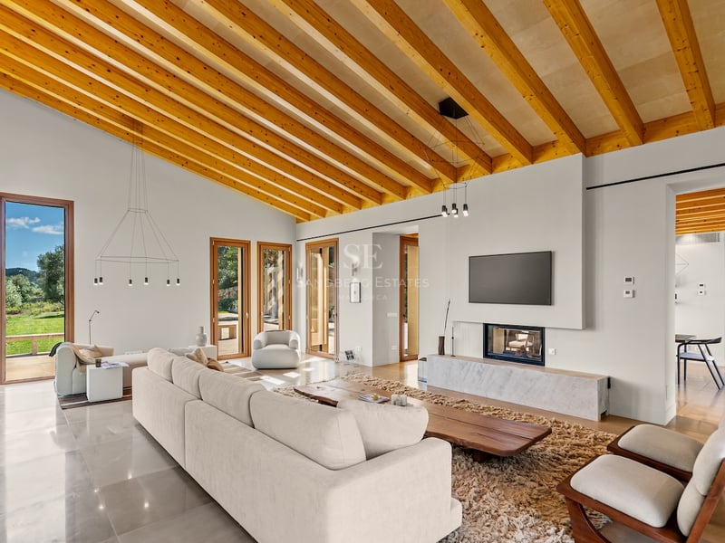 Grand salon moderne avec hauts plafonds à poutres, grand canapé, cheminée et vue sur le jardin.