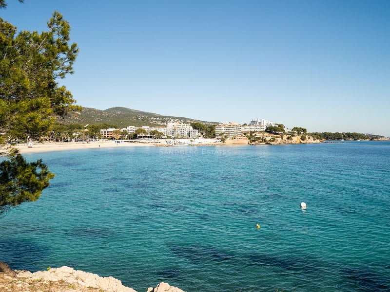 Weitläufiger Blick auf eine mediterrane Bucht mit klarem türkisfarbenem Wasser, Sandstrand und weißen Gebäuden.