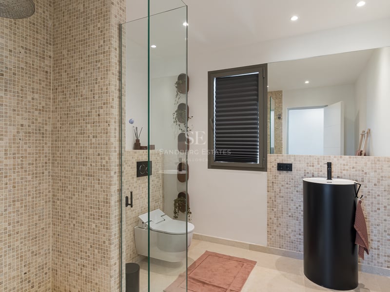 Modernt badrum med mosaikkakel, duschvägg i glas, mattsvart pelarhandfat och vägghängd toalett.