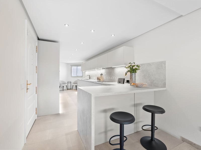 Une cuisine blanche moderne avec un comptoir en pierre, deux tabourets noirs et un éclairage LED intégré.