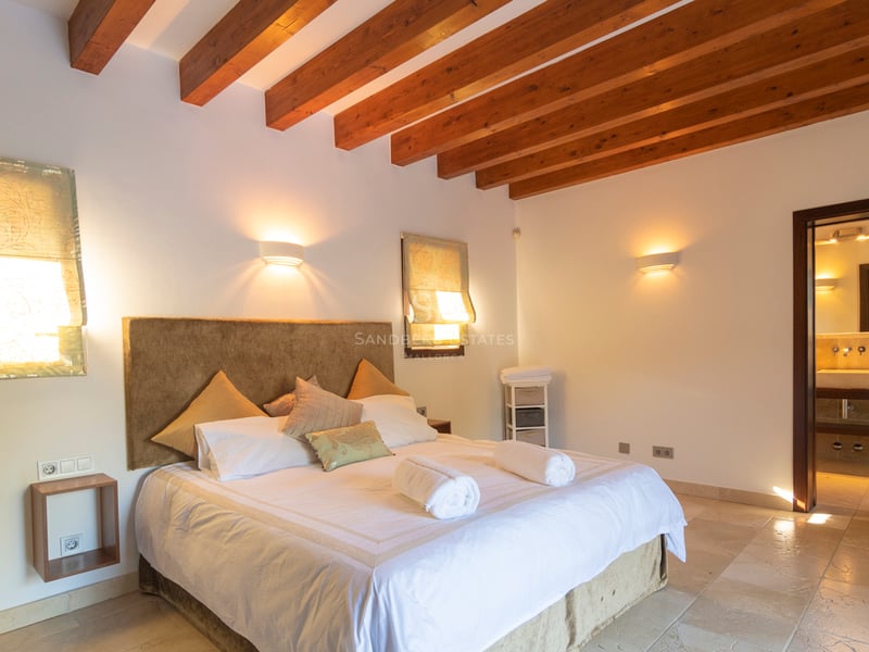 Sovrum med mörka takbjälkar i trä, stengolv och en öppen dörr till ett privat badrum.