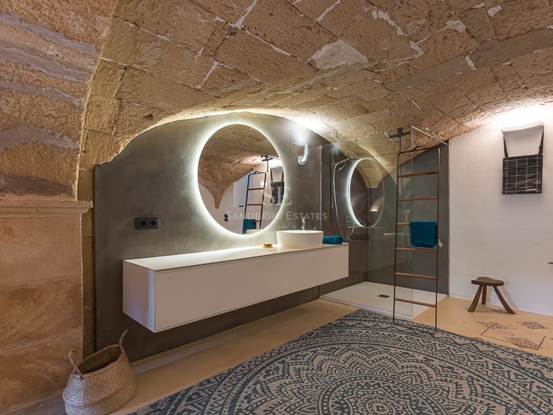 Salle de bain avec plafond voûté en pierre, miroir rétroéclairé et meuble vasque suspendu blanc.