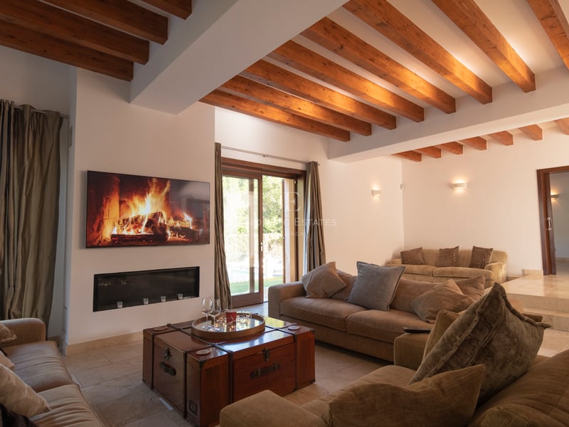Vardagsrum med takbjälkar i trä, modern öppen spis, stengolv och stora glasdörrar mot trädgården.
