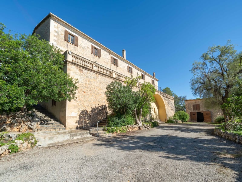 Grande villa en pierre avec terrasse surélevée, volets en bois et arbres sous un ciel bleu clair.
