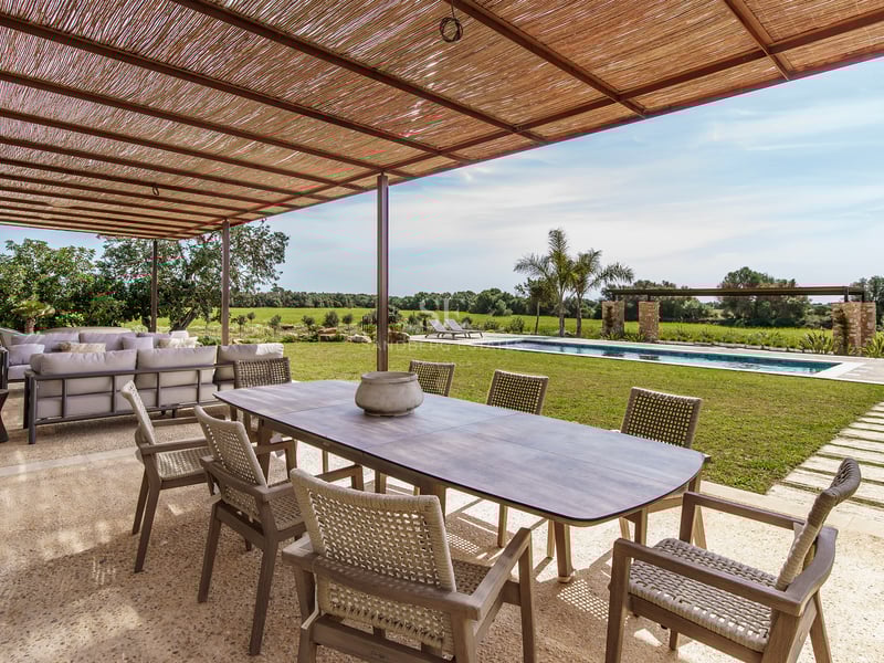 Großer Holzesstisch unter einer Schilfpergola auf einer Steinterrasse mit Blick auf Pool und Rasen.