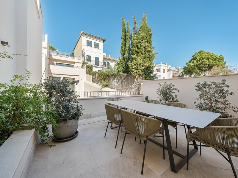 A contemporary outdoor terrace featuring a large dining table with six woven chairs on a stone-tiled floor.