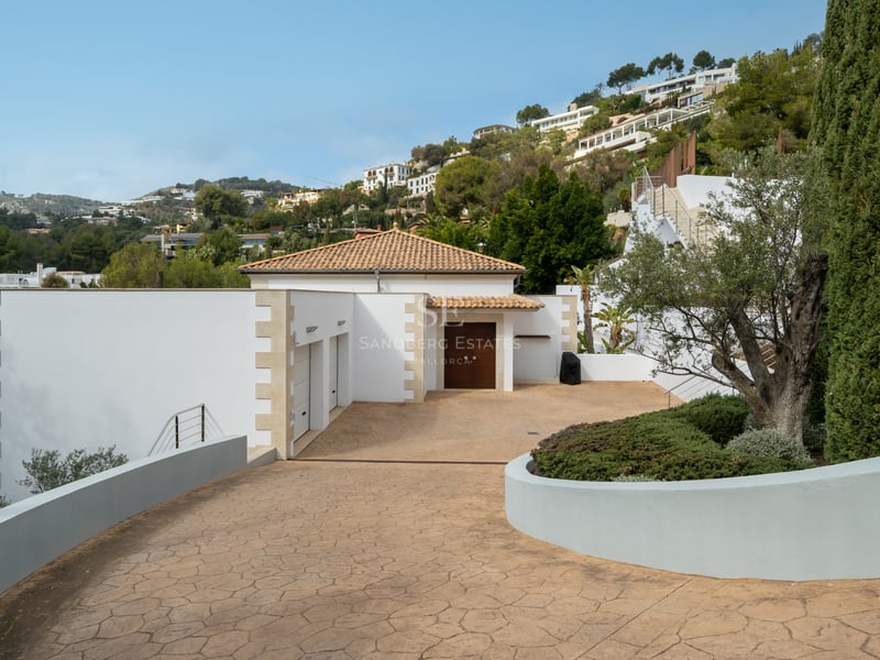Une allée privée pavée menant à une villa blanche avec toit en terre cuite, double garage et végétation luxuriante.