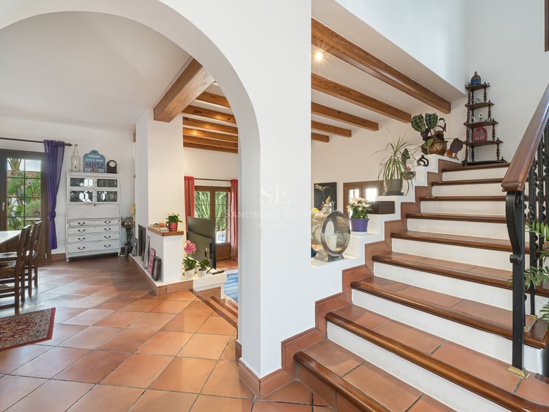 Interior view showing terracotta floors, an arched entrance, wooden ceiling beams, and a staircase with iron railings.