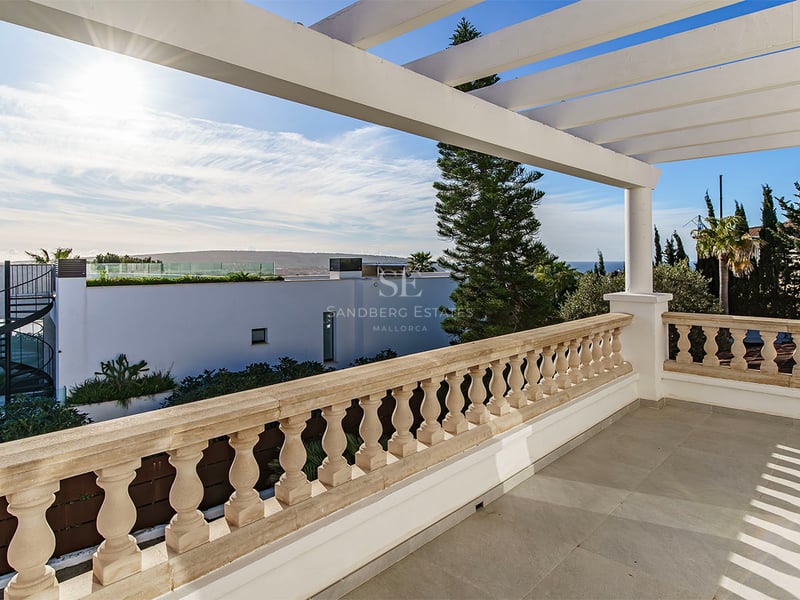 Rymlig terrass med stenbalustrad och vit pergola med utsikt över kustlandskapet och en klarblå himmel.