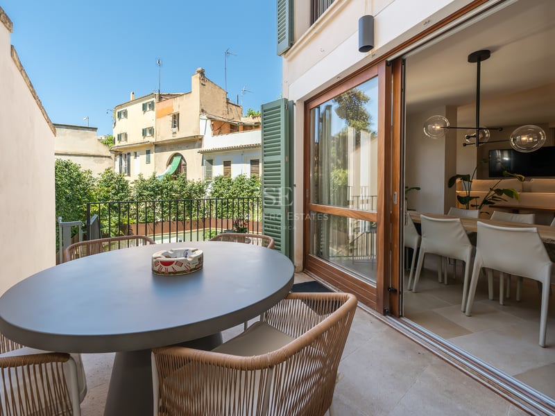 Terrasse avec table ronde grise, chaises en corde tressée et baies vitrées ouvrant sur le séjour.