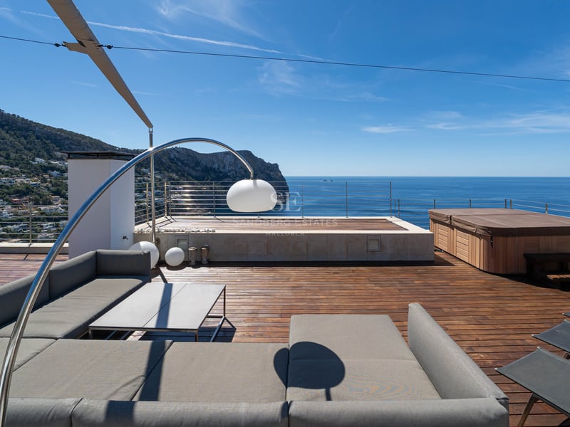 Modern rooftop terrace with grey outdoor sofa, wooden decking, and a hot tub overlooking the blue sea and mountains.