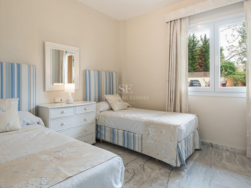Sunlight-filled twin bedroom featuring blue and white linens, white dresser, and polished marble floors.