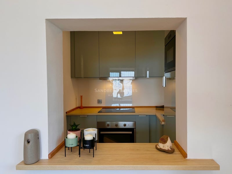 View through a wall opening of a modern kitchen with sage green cabinets and wood countertops.