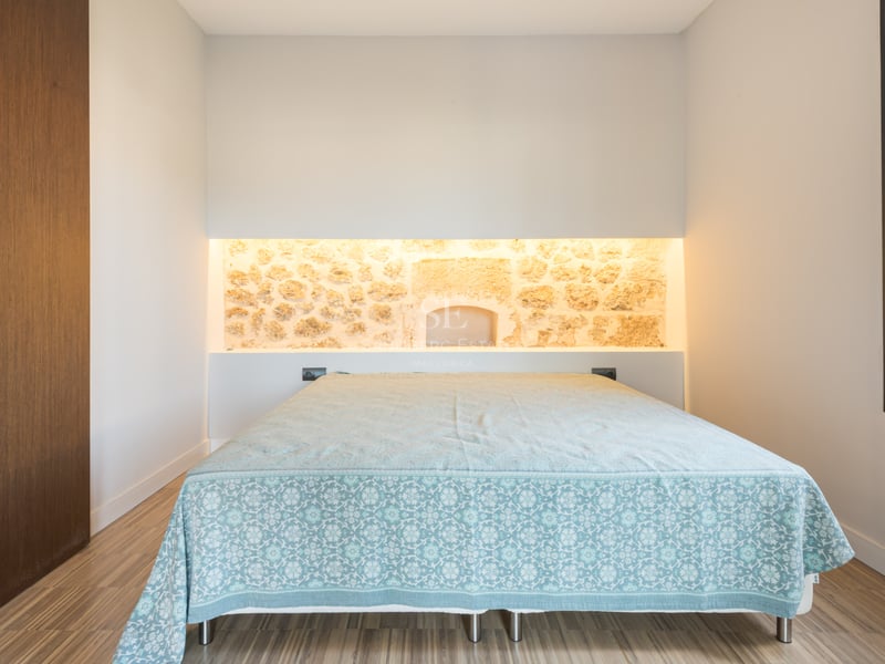 Sovrum med en säng, en belyst stenvägg i bakgrunden och ljust trägolv.