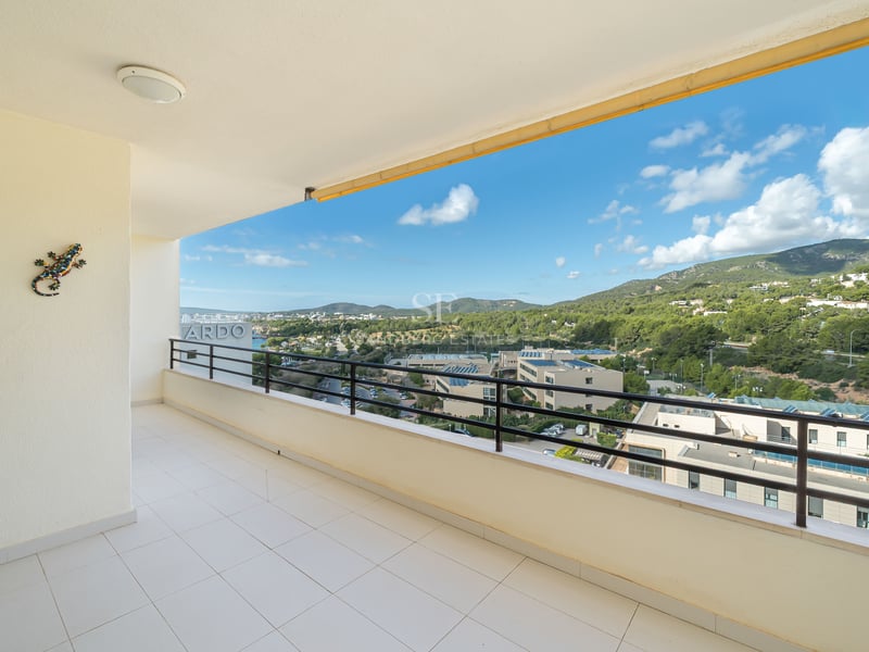 Spacious white tiled balcony with a black metal railing overlooking green hills and a blue sky with white clouds.