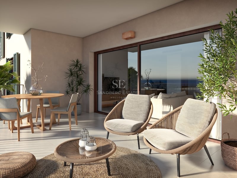 Spacious covered terrace featuring wicker armchairs, a wooden dining table, and large glass doors overlooking the sea.