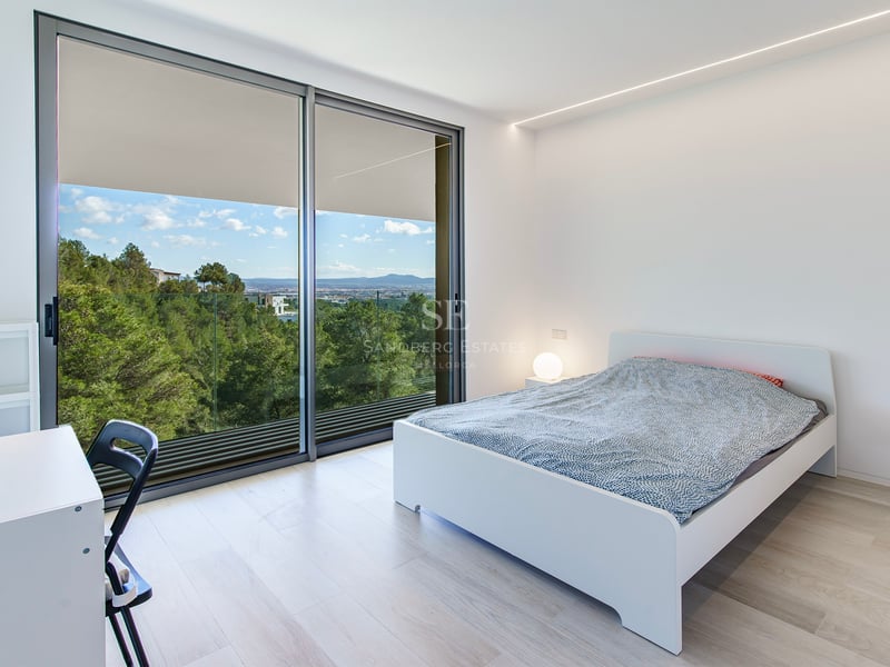 Chambre lumineuse avec lit blanc, parquet clair et grandes baies vitrées donnant sur une forêt et des montagnes.