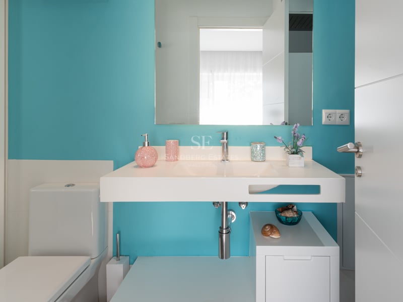 Salle de bain moderne avec mur turquoise, vasque suspendue blanche, grand miroir et toilettes blanches.