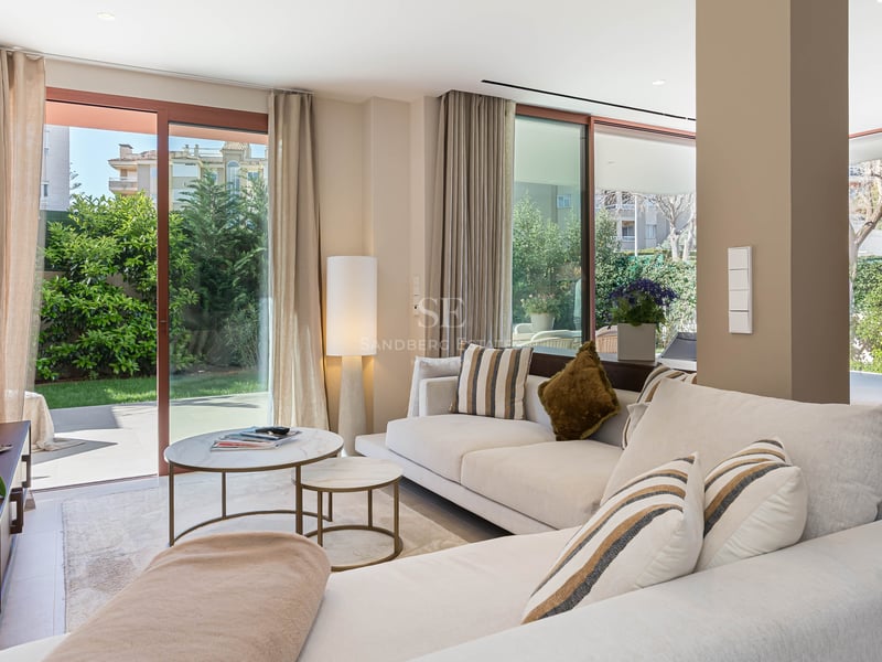 Bright modern living room with cream sofa, marble tables, and floor-to-ceiling glass doors opening to a lush garden.