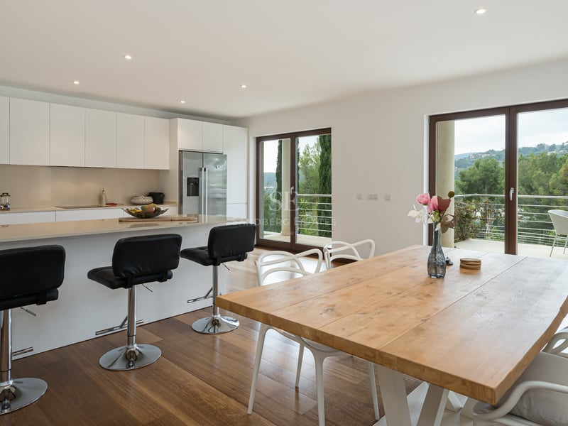 Cuisine contemporaine blanche avec îlot central, table de repas en bois et baies vitrées ouvrant sur la nature.