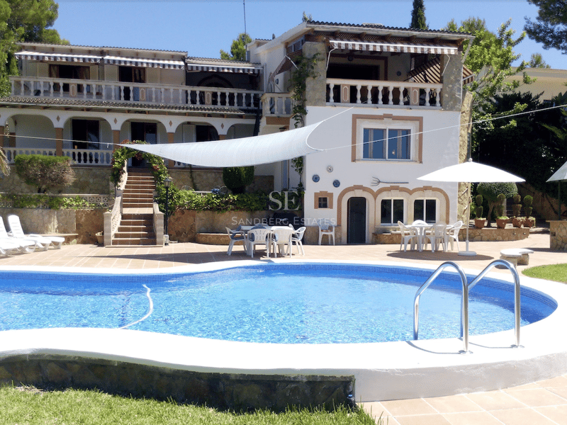 Stor kurvad pool framför en vit medelhavsvilla i flera plan med terrasser i terracotta och stenmurar.