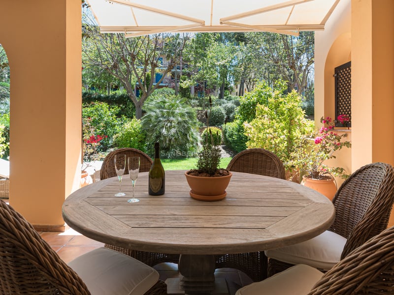 A covered terrace with a round wooden table, wicker chairs, and a view of a lush Mediterranean garden.