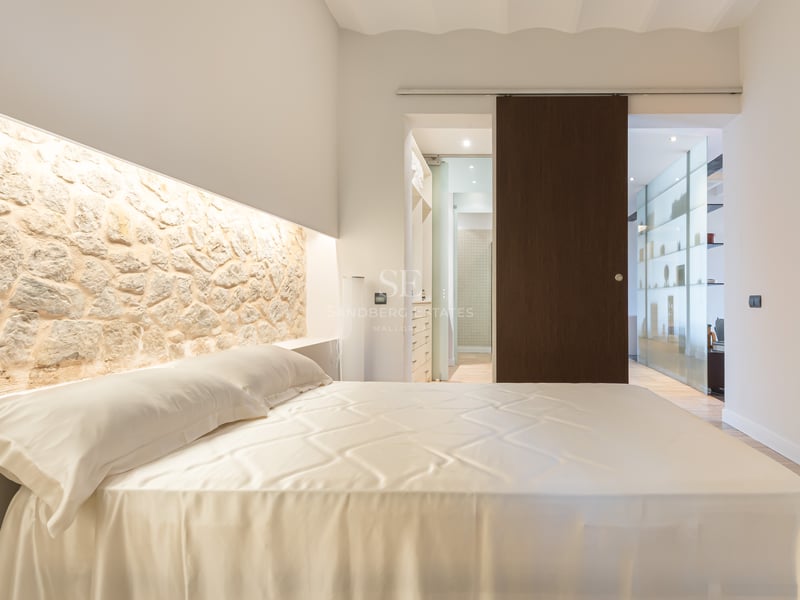 Lyxigt sovrum med en stor säng, en belyst fondvägg i natursten och en skjutdörr i mörkt trä.