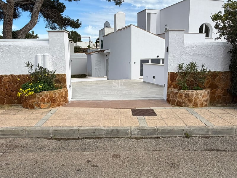 Entrée d'une villa blanche avec murs en pierre, jardinières de fleurs jaunes et allée pavée sous des pins.