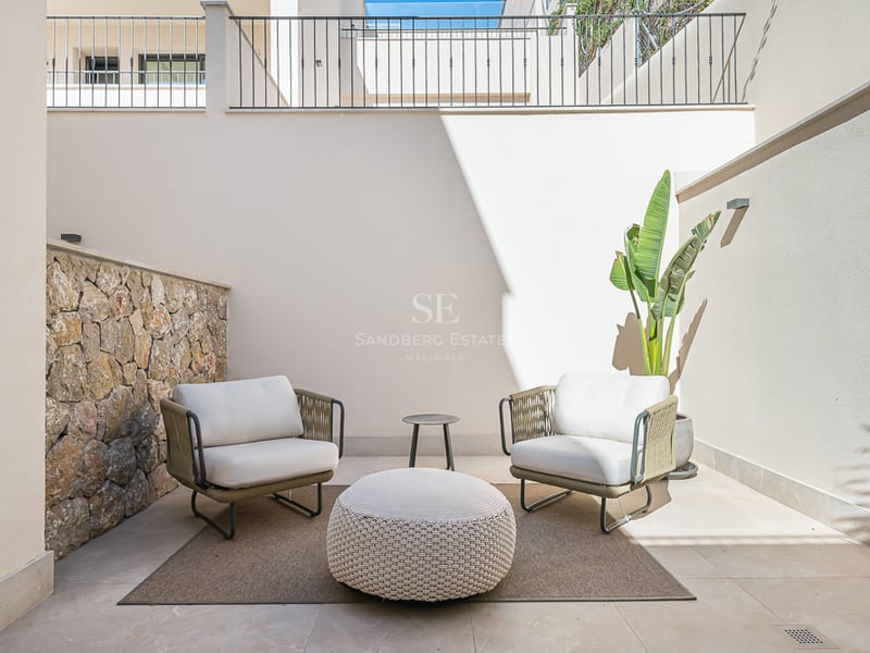 A sunlit private patio featuring a natural stone wall, two modern armchairs, a large woven pouf, and a potted plant.