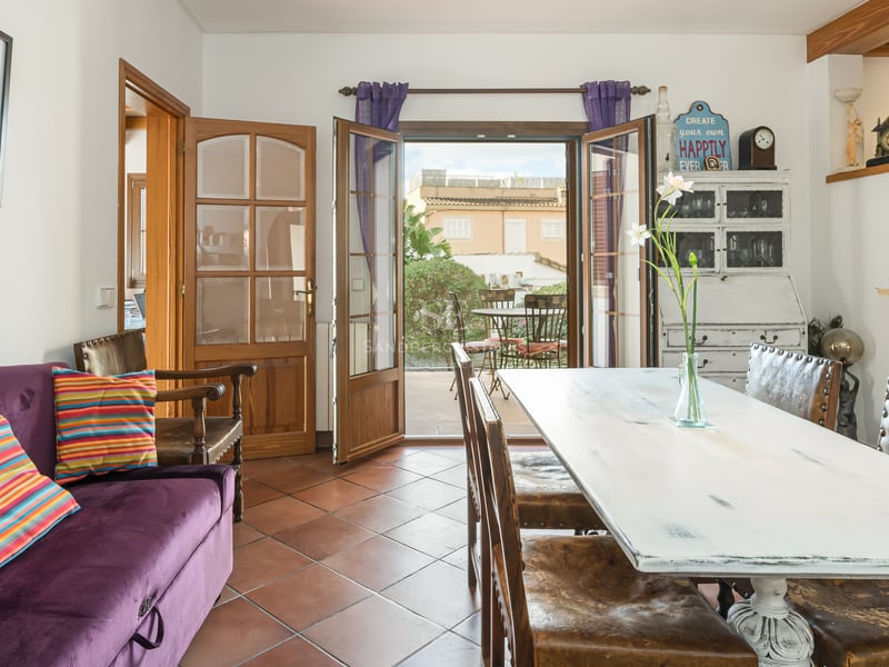 Interior dining room featuring terracotta tile floors, a rustic white wooden table, and open doors leading to a sunny patio.