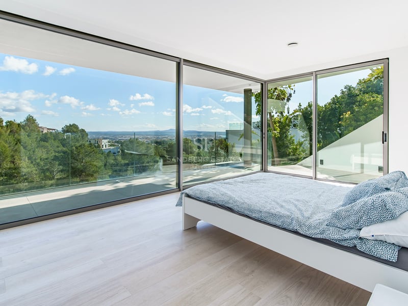 Chambre moderne avec lit blanc et grandes baies vitrées donnant sur une forêt verdoyante et la ville au loin.