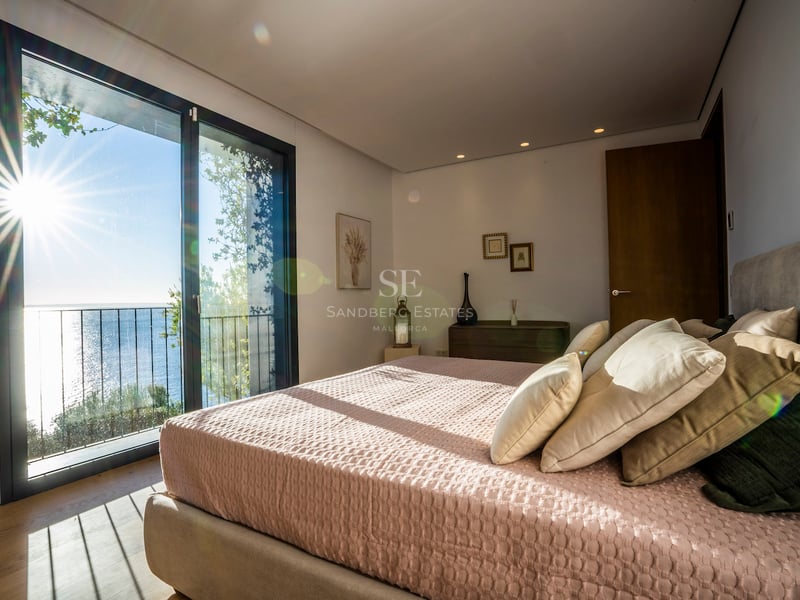 Helles Schlafzimmer mit großem Bett, rosa Bettwäsche, Eichenparkett und bodentiefem Fenster mit Blick auf das blaue Meer.