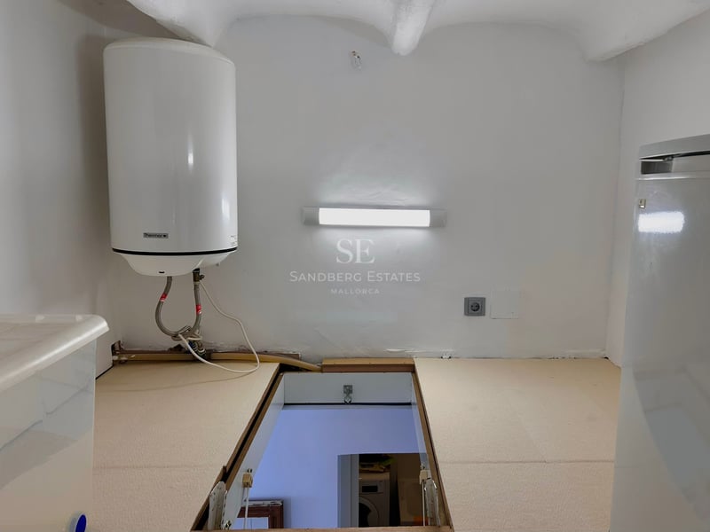 White utility room with an electric water heater on the wall and a rectangular hatch open to the floor below.
