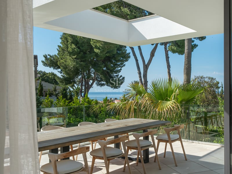 Espace repas extérieur moderne sur une terrasse avec table en bois, garde-corps en verre et vue sur la mer.