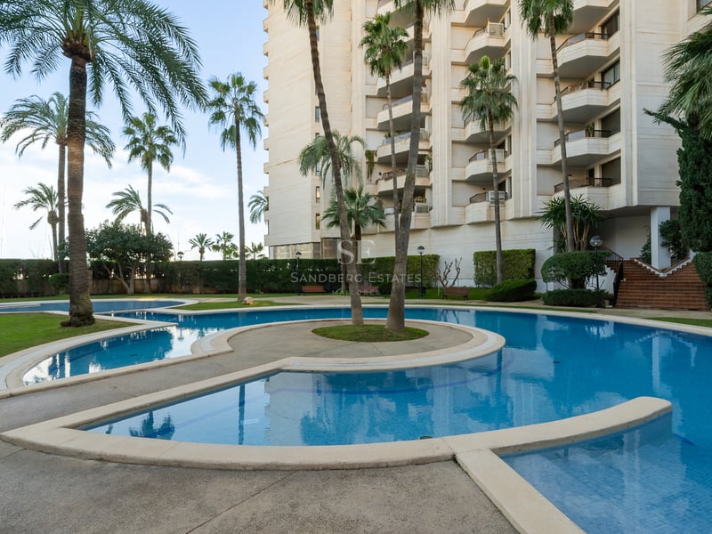 Stor svängd pool omgiven av höga palmer och ett lägenhetshus under en klar himmel.