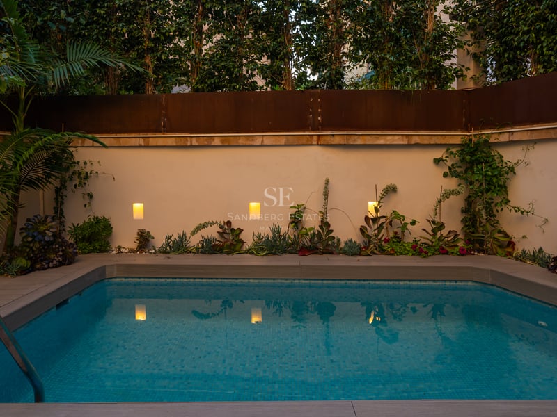 Piscine rectangulaire moderne au crépuscule avec eau turquoise, appliques murales chaleureuses et végétation tropicale.