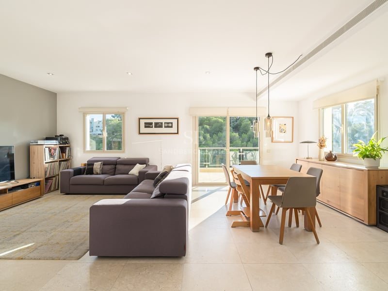 Ljust vardagsrum med grå soffa, matbord i trä och stora fönster som vetter mot en skog.