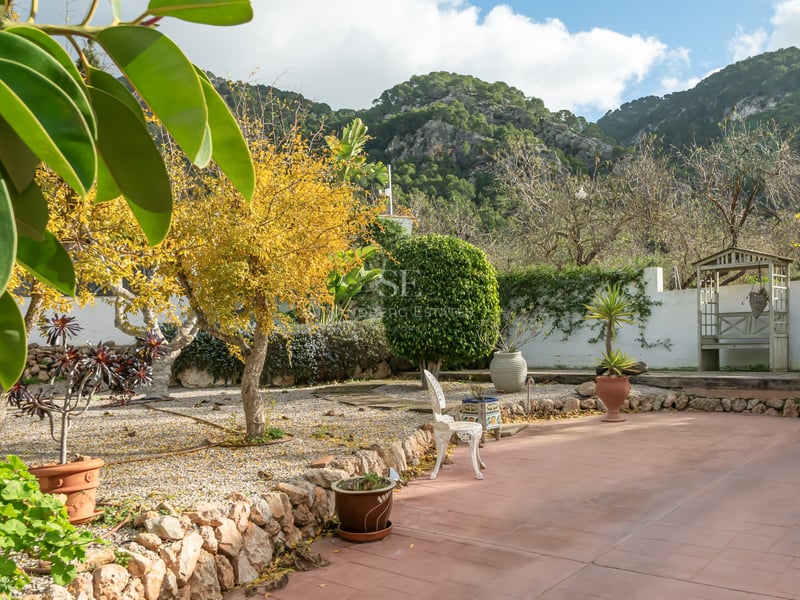 Terracotta terrace and landscaped garden with stone walls and mature trees against a rocky mountain backdrop.