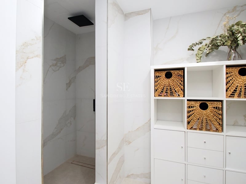 Ett modernt badrum med en rymlig dusch, väggar i marmoreffekt, svart takdusch och vit hylla med korgar.