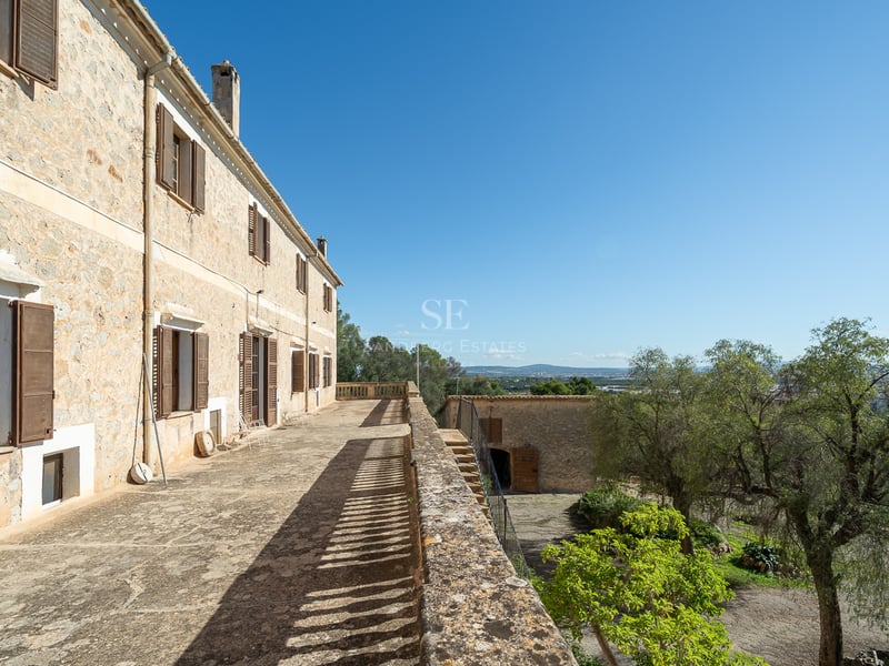 Longue terrasse en pierre sur un bâtiment rustique avec volets en bois, surplombant un paysage verdoyant.