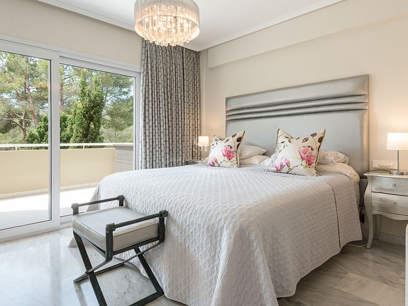 Bright master bedroom with marble floors, silver headboard, and sliding glass doors opening to a terrace with tree views.