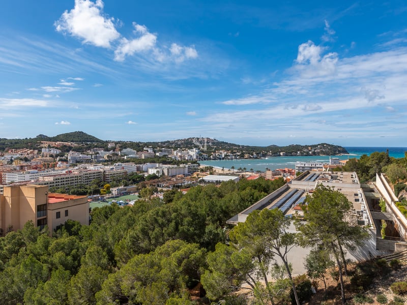 Vue surélevée d'une ville côtière méditerranéenne avec mer bleue, pinèdes et montagnes sous un ciel clair.