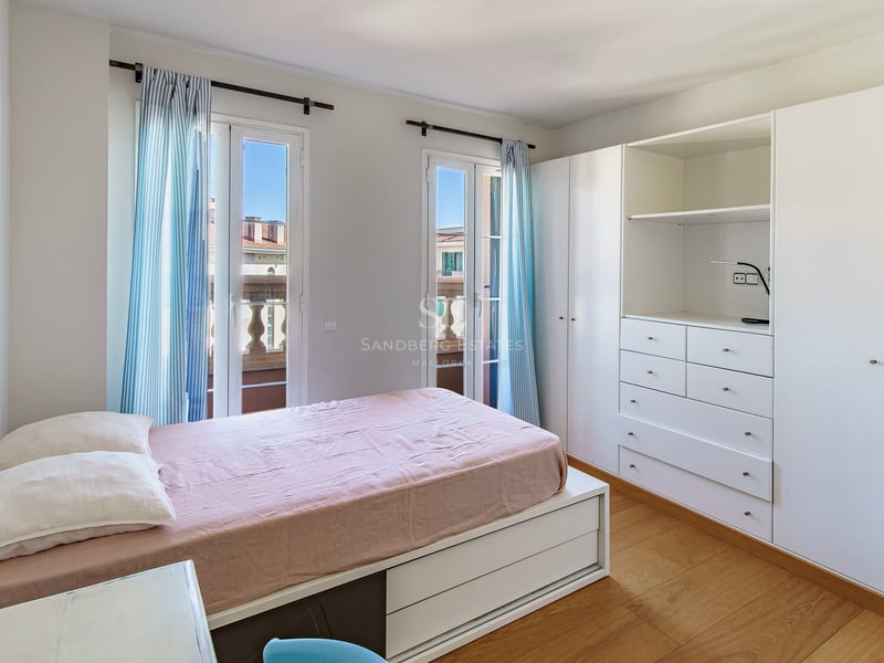 Ljust sovrum med rosa sängkläder, vita garderober och franska dörrar med blårandiga gardiner.